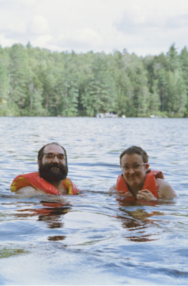 A photograph of two people from the shoulders up, both wearing orange life vests and glasses, floating in a lake with green trees in the background. Joe is on the left, Sam on the right.